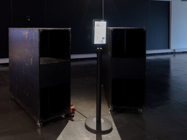 A dimly lit exhibition space featuring two large, black, worn wooden speaker cabinets on casters, positioned on either side of a tall, black stand. A small illuminated screen is mounted on the stand, displaying text and a logo. The speakers and screen are the focal points, with a dark background and a reflective floor adding to the atmosphere.