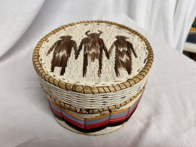 Round quillwork basket with lid featuring three human-like figures, likely shamans, and colorful bands woven around the sides.