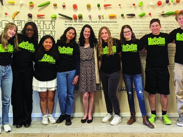 A diverse group of MSU students stand with their supervisor, all students wearing black t-shirts with the MSU Museum logo in lime green. Their supervisor, Caroline White, stands in the middle in a patterned dress. Everyone smiles at the camera, posing in a front of a lead exhibit wall decorated with rows of hanging plastic fruit above their heads.