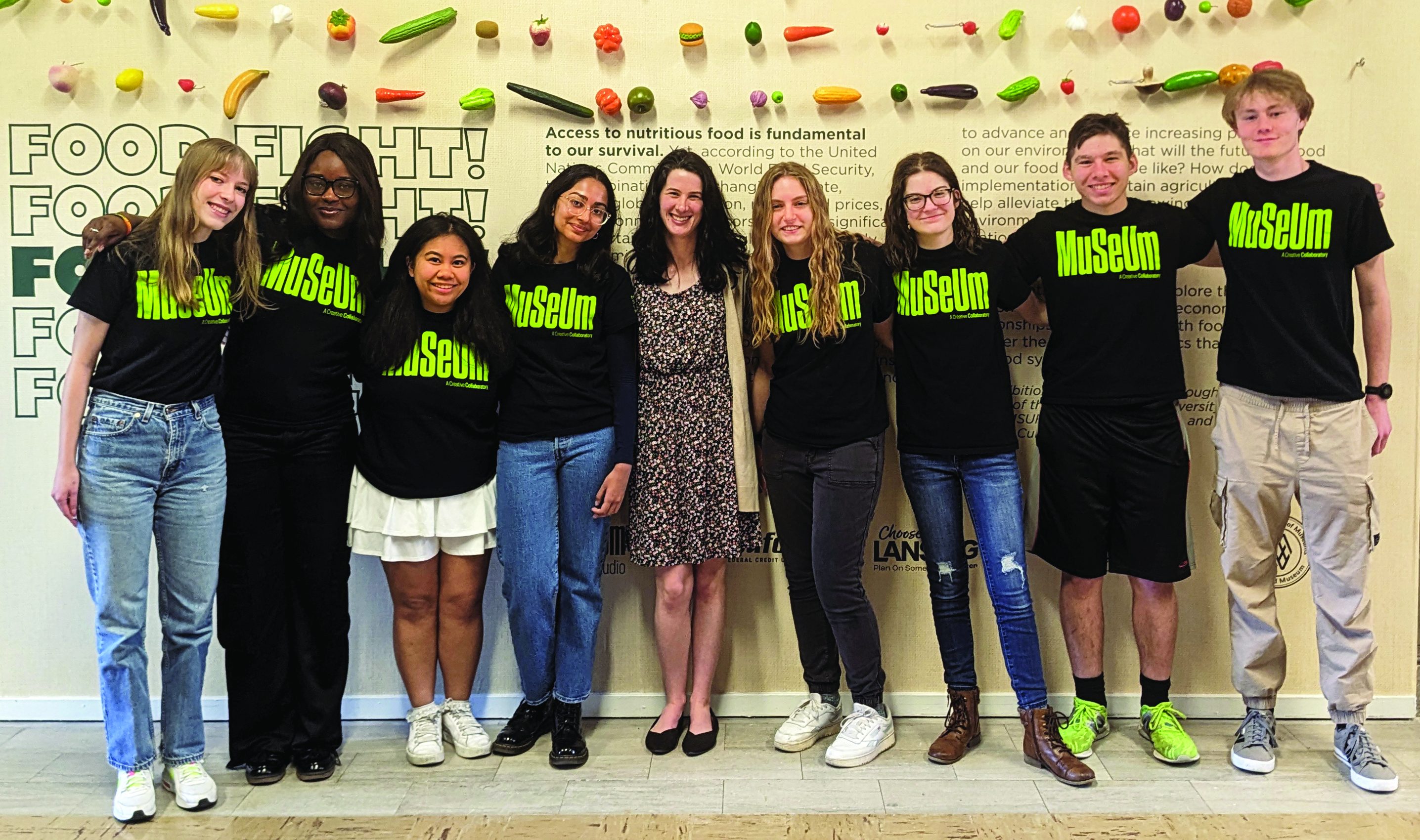 A diverse group of MSU students stand with their supervisor, all students wearing black t-shirts with the MSU Museum logo in lime green. Their supervisor, Caroline White, stands in the middle in a patterned dress. Everyone smiles at the camera, posing in a front of a lead exhibit wall decorated with rows of hanging plastic fruit above their heads.