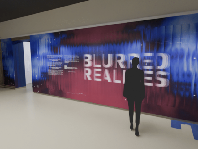 A person stands in front of a large, colorful wall display titled "Blurred Realities" in bold white text. The background features a digital, glitch-inspired design in shades of blue, purple, and red. Informational text panels are arranged across the wall, with some content also displayed on the floor near the viewer. The setting appears to be a modern exhibition or gallery space.