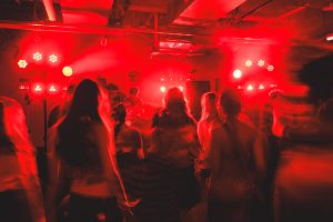 A crowd of people dances under intense red lighting in a dark indoor venue, facing a DJ performing on stage. The figures appear blurred due to motion, conveying the energy and movement of the scene.