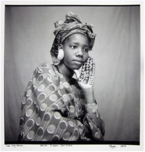 Black-and-white portrait of a young woman in patterned traditional attire and headwrap, wearing large textured earrings. She rests her face on her hand, which displays geometric henna designs.