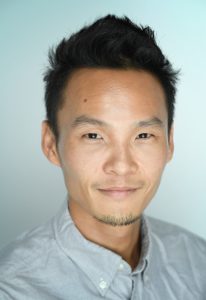 ALT text: Close-up portrait of a man with short, spiked black hair, a trimmed goatee, and a slight smile, wearing a light gray button-up shirt against a pale blue background.