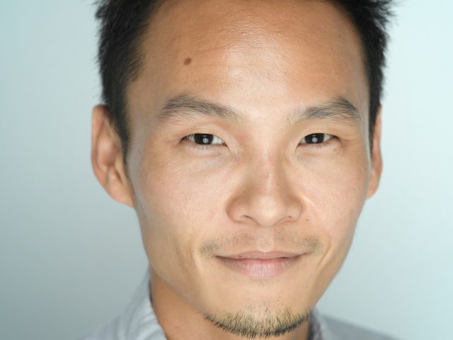ALT text: Close-up portrait of a man with short, spiked black hair, a trimmed goatee, and a slight smile, wearing a light gray button-up shirt against a pale blue background.