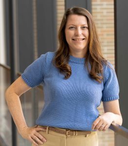 Woman with long brown hair wearing a short-sleeved blue knit top and beige pants, standing indoors near a glass railing, smiling at the camera.