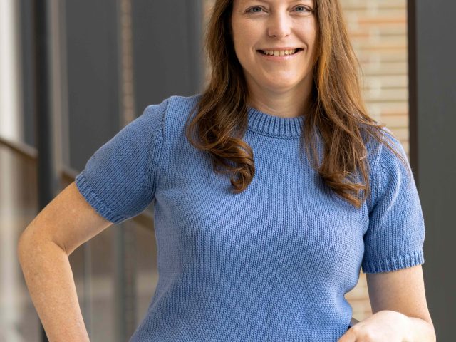 Woman with long brown hair wearing a short-sleeved blue knit top and beige pants, standing indoors near a glass railing, smiling at the camera.
