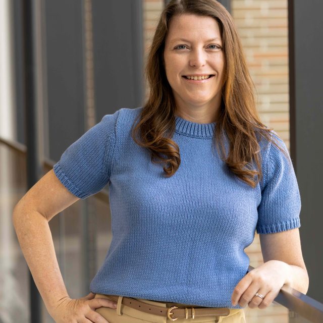 Woman with long brown hair wearing a short-sleeved blue knit top and beige pants, standing indoors near a glass railing, smiling at the camera.
