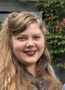 Close-up portrait of a woman with long, wavy light brown hair and red lipstick, smiling at the camera. She is wearing a gray collared shirt and hoop earrings, standing in front of a dark wooden wall with green and red ivy leaves.