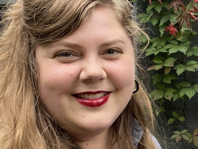 Close-up portrait of a woman with long, wavy light brown hair and red lipstick, smiling at the camera. She is wearing a gray collared shirt and hoop earrings, standing in front of a dark wooden wall with green and red ivy leaves.