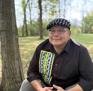 A smiling person wearing glasses, a black and white patterned flat cap, and a black shirt with a colorful, geometric ribbon applique sits outdoors near a tree. The background shows a wooded area with green grass and tall trees under a bright sky.