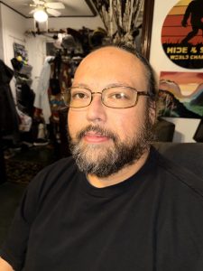 A man with glasses, a full beard, and long hair tied back is seated indoors, facing the camera under warm lighting. He is wearing a black shirt. The background includes a cluttered room with hanging clothes, a ceiling fan with a light, and a circular wall sign featuring a Bigfoot silhouette and the words "Hide & Seek World Champion.