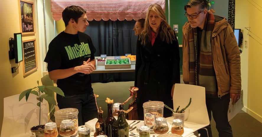 In a colorful exhibit space painted bright yellow and green, an MSU student working as an MSU Museum CoLaborator explains the exhibit to two visitors dressed in winter coats. In front of the visitors sits a table spread with different cutlery and containers full of natural decomposition materials such as mushrooms and vermicompost.