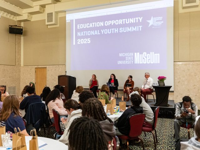A panel of five speakers sits on a stage in front of a large screen displaying "Education Opportunity National Youth Summit 2025" at Michigan State University Museum. The audience, composed mostly of young people, is seated at round tables in a conference room, some listening attentively while others look at their phones or talk.
