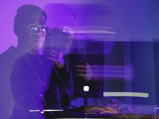 DJ Carl Craig wearing sunglasses and a chain performs under purple and blue lighting, with motion blur and double exposure effects creating a dynamic, layered visual.