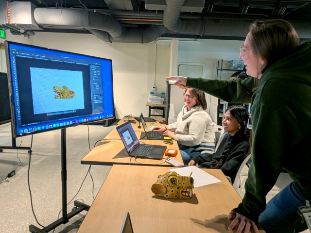 MSU Museum employees smile as they work on digitizing a jaguar mask in the Bloomberg lab. Closest to the camera, Carrie Walker, with long brunette hair and wearing an MSU sweatshirt, points at a large computer screen on a rolling stand while smiling. On the screen, Photoshop is open to show a side profile of the jaguar mask which rests on the table by Carrie's hand. Next to her, a student worker with black hair in a ponytail wearing a black sweatshirt smiles. A similar picture is up on the laptop in front of her. Further down the row of tables, Curator Samantha Ellens, with short brunette hair and glasses, wearing a grey turtleneck sweater, smiles as she also works on a laptop. The scene around them is an office space with visible ducts and piping, white walls, and storage spaces.