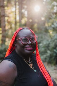 A person with long, bright red braids and large round pink glasses smiles while standing outdoors in a forested area. They wear a black sleeveless top, gold necklaces, and have facial piercings and visible tattoos. Sunlight filters through the trees in the background.