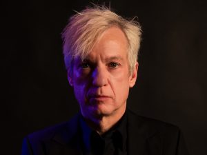 Close-up portrait of Chris Vrenna with short white hair wearing a black suit and black shirt, looking directly at the camera with a serious expression. Blue and warm orange lighting cast contrasting tones across their face against a dark background.