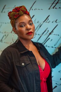 Mahogany Brown wearing a red-and-yellow patterned headwrap, large hoop earrings, a black denim jacket, and a red V-neck top stands in front of a light blue wall with cursive writing. She faces the camera with a neutral, confident expression, her arm raised slightly as if resting against the wall.