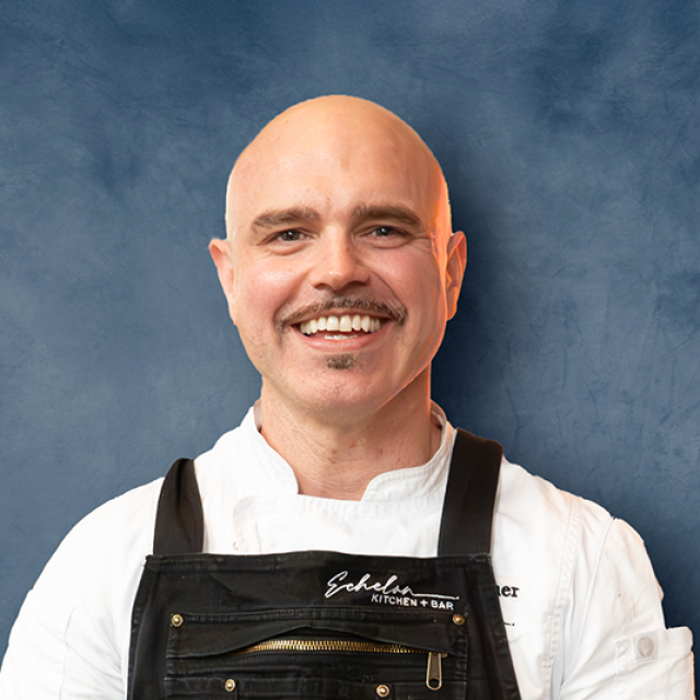 Block image Smiling bald man with a mustache wearing a white chef's coat and black apron embroidered with 