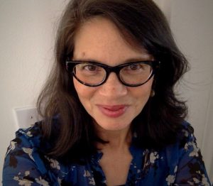 A woman with dark, shoulder-length hair and black cat-eye glasses smiles at the camera. She is wearing a blue floral blouse and is standing indoors against a light-colored wall.