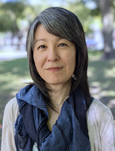 A woman with straight, shoulder-length salt-and-pepper hair stands outdoors in a shaded park. She wears a light-colored cardigan over a white top and a dark blue scarf. The background shows grass and trees softly out of focus.