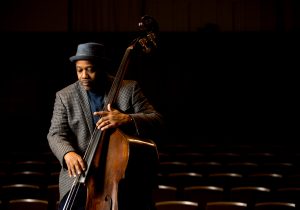 A man wearing a hat and checkered blazer plays an upright bass in a dimly lit auditorium with empty rows of chairs behind him. He appears focused, with his eyes downcast and his fingers positioned on the strings.