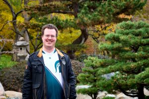 Ryan Thompson wearing glasses, a dark jacket, and a teal-and-white shirt stands in a landscaped garden with evergreen trees and yellow autumn foliage. A stone lantern and sculpted branches appear in the background.