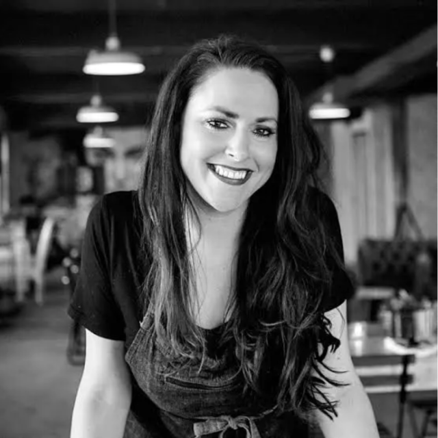 Block image Smiling woman with long dark hair wearing a dark T-shirt and apron, leaning forward slightly in a warmly lit restaurant interior; image is in black and white.