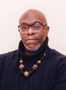A bald man with glasses and a short gray beard wears a dark blue cable-knit turtleneck sweater and a chunky beaded necklace in shades of brown and black. He looks directly at the camera against a plain light background.