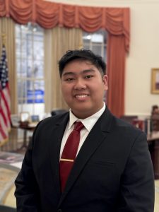 Young man in a black suit and red tie smiles at the camera while standing in a formal office resembling the Oval Office, with an American flag, desk, and draped windows visible in the background.