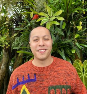 A person with a shaved head and a warm smile stands in front of lush, tropical foliage. They are wearing an orange knit sweater with colorful geometric patterns.