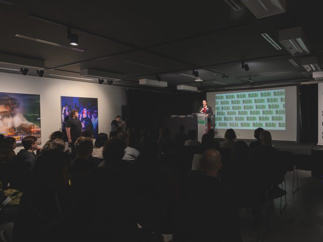 View from the audience in the Commons. Many people sit in black chairs arranged in rows in front of a stage where a woman in a burgundy dress speaks at a podium.
