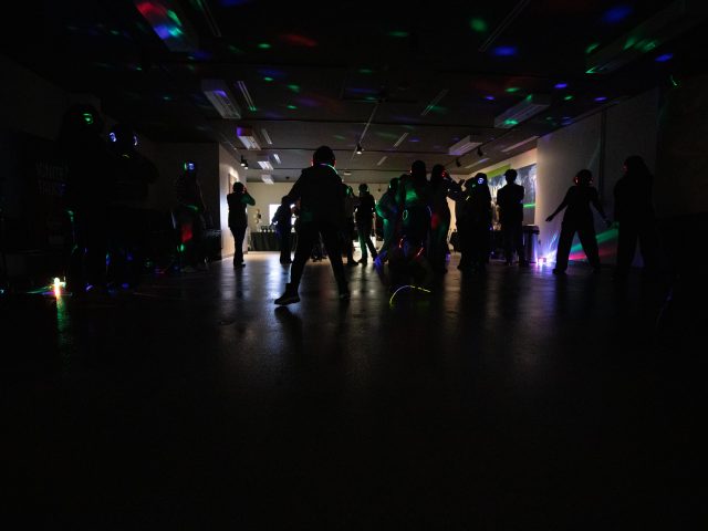 A dimly lit space filled with students dancing while wearing glowing headphones. Disco lights bounce off the walls to set the mood.