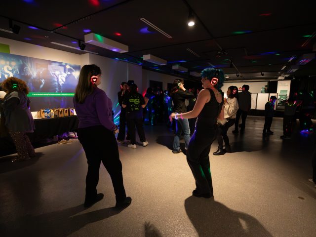 A large empty space where students can grab food at a nearby table in between dancing. Centered in the image, two students wearing glowing headphones dance while other students can be seen doing the same in the background.