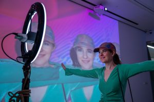 Woman wearing a green long-sleeve top and multicolored cap stands with arms outstretched in front of a projected image of her face, illuminated by purple and blue lighting, with a ring light and projector visible in a studio setting.