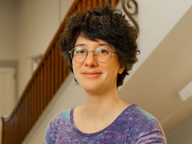 Block image A woman with very short brown hair and glasses smi.es at the camera, wearing a blue/purple blouse and posing in front of a staircase