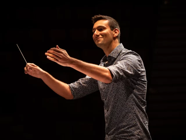 Block image A man wearing a blue button up holds his hands up, conducting an orchestra not in the photo. Background is black.