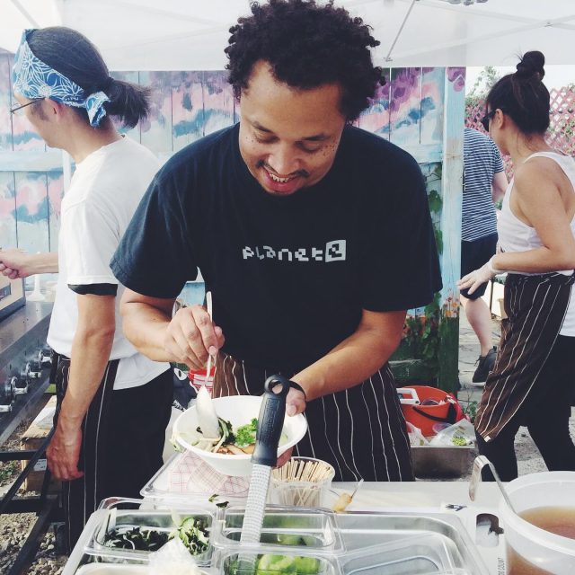 Block image Man with short curly hair wearing a dark T-shirt and black apron, standing outdoors bent over a serving container of food. Other chefs are visible in the background.