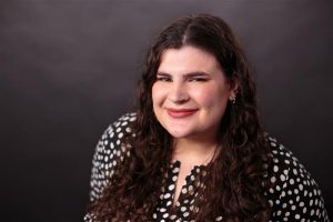 Head-and-shoulders studio portrait of a woman with long, dark curly hair smiling at the camera against a dark gray background, wearing a black blouse with white polka dots and small hoop earrings.