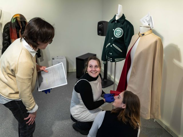 Three MSU students examine clothing on mannequins in a studio or exhibit space; one holds a printed document while two others adjust a beige cape with a red lining, smiling and conversing.