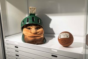 A vintage Sparty head wearing a green helmet with a plume sits on a white shelf next to an old leather basketball labeled with a 1951 game score.