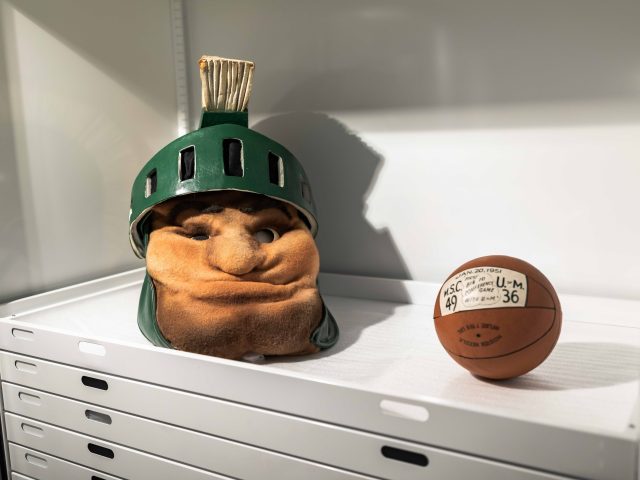A vintage Sparty head wearing a green helmet with a plume sits on a white shelf next to an old leather basketball labeled with a 1951 game score.