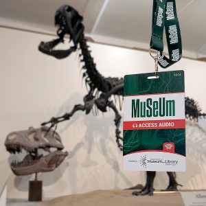 A museum access badge labeled “MuSeum – Access Audio” hangs in the foreground, with a blurred dinosaur skeleton exhibit and skull visible in the background.