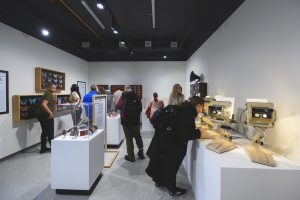 Visitors explore a museum gallery, viewing displays of vintage electronic devices and looking through interactive viewing stations mounted on a white wall.