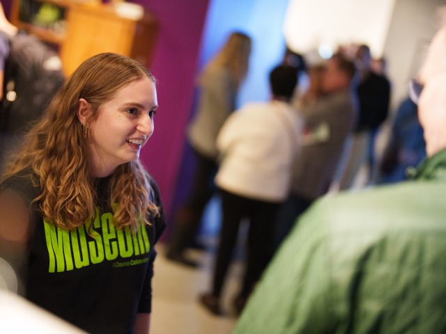 MSU student CoLaborator Rachel, with long wavy blonde hair and wearing a black MSU Museum shirt, chats with guests at an exhibit opening. Various visitors can be seen out of focus around her.