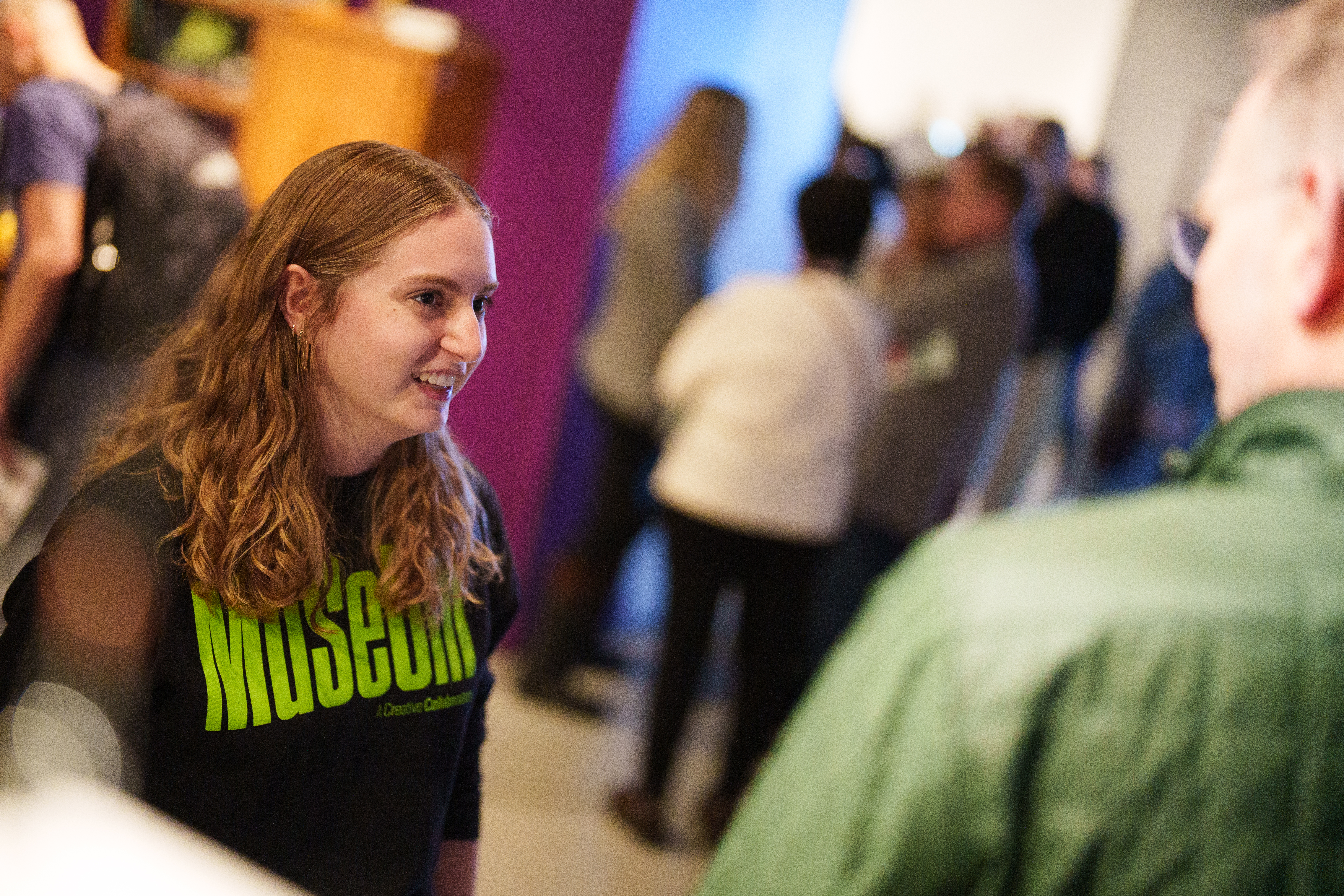 MSU student CoLaborator Rachel, with long wavy blonde hair and wearing a black MSU Museum shirt, chats with guests at an exhibit opening. Various visitors can be seen out of focus around her.