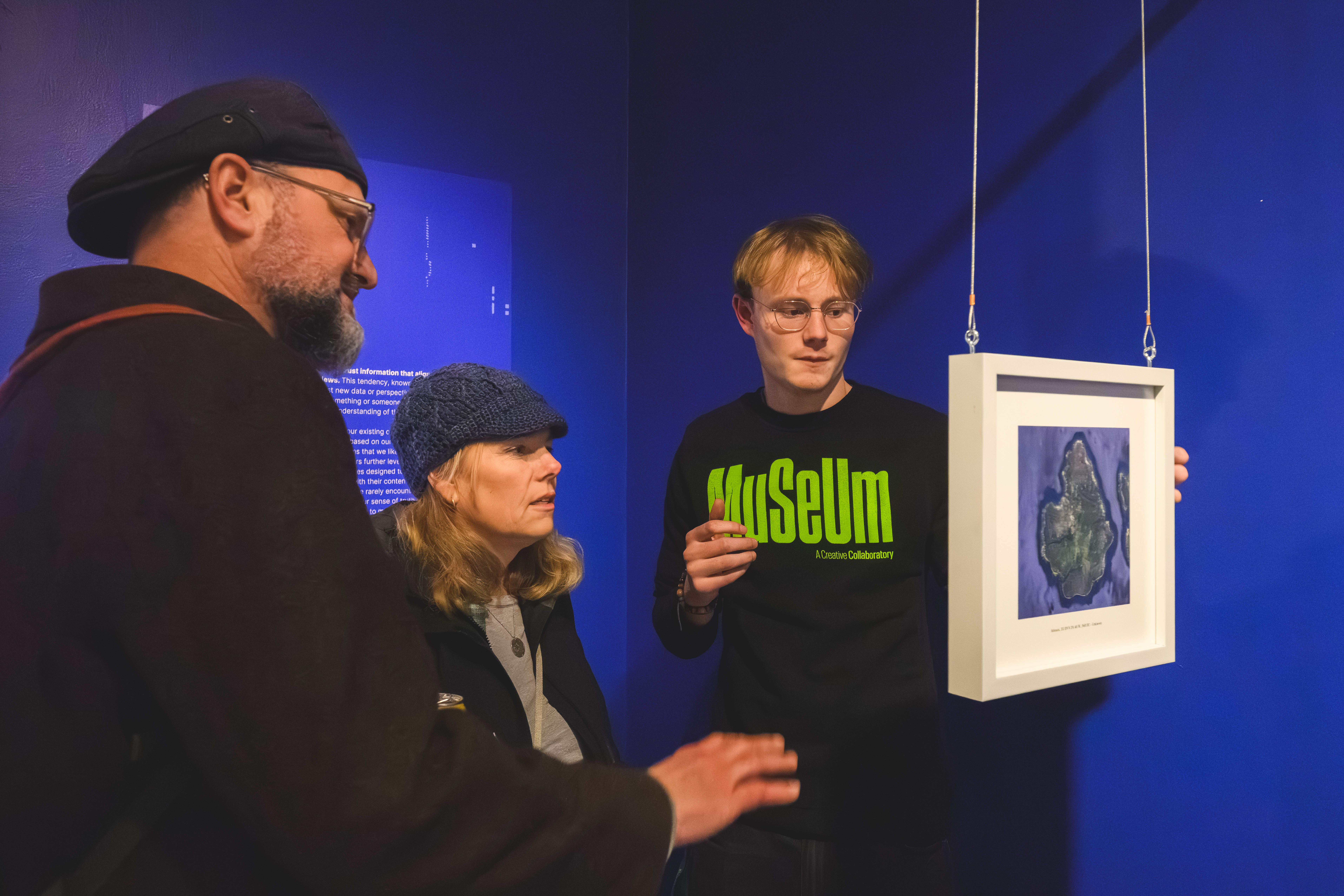 MSU Student CoLaborator Sam Booth, wearing glasses and a black MSU Museum shirt, explains an exhibit to two visitors. Part of the exhibit includes a small white frame suspended from the ceiling containing an image of an AI-generated map.