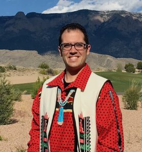 Smiling person wearing glasses and a red patterned shirt with a cream-colored vest and beaded necklace, standing outdoors in a desert landscape with mountains and a partly cloudy sky in the background.