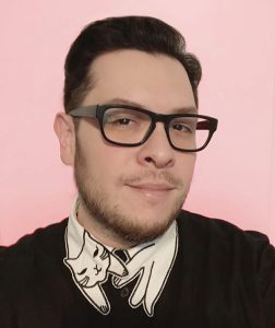 Close-up portrait of a person with short dark hair and black rectangular glasses, wearing a black sweater over a white collared shirt with a cat illustration on the collar, set against a solid pink background.